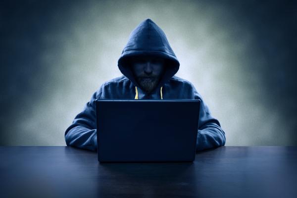 A man whose face is obscured by a hoodie typing on a laptop in front of him