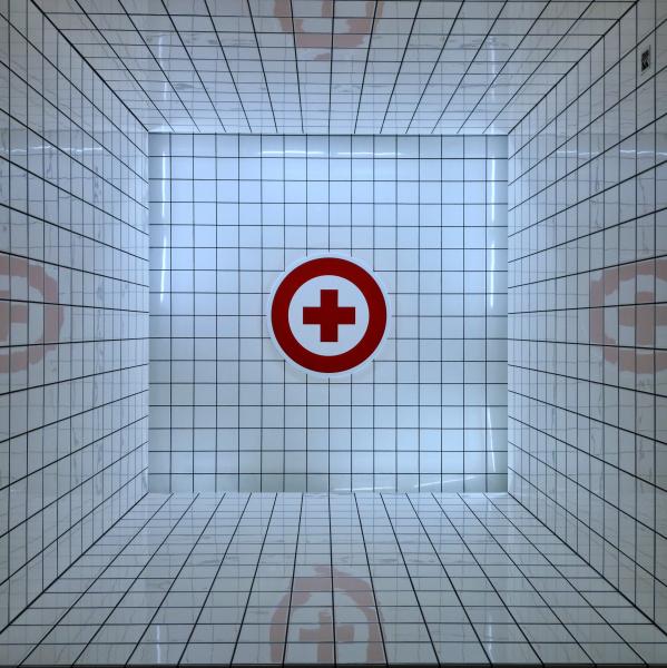 A white tiled corridor, at the end of the corridor is the universal symbol for healthcare, a red cross surrounded by a red circle. Photo by Enric Moreu on Unsplash