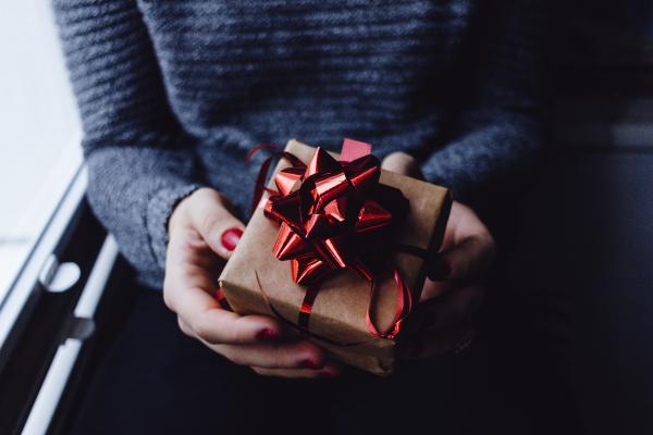 A brown paper wrapped present, with a shiney red bow being held by a pair of hands with red nail polish. Photo by freestocks on Unsplash