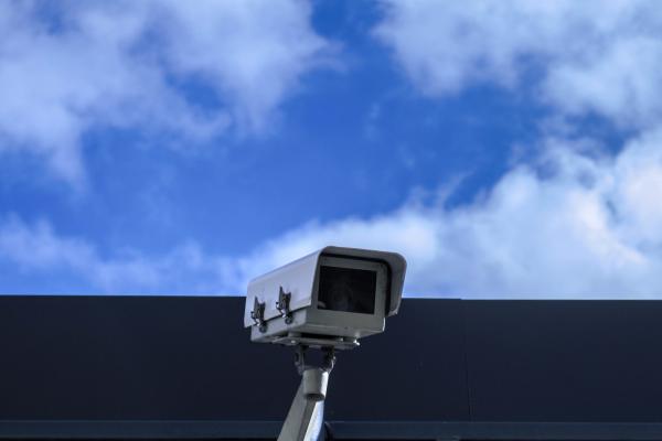 Image of a CCTV Camera imposed against a rooftop and a blue sky. Photo by Nathy dog on Unsplash